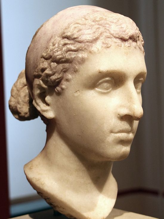Marble head of a girl. She has similar features to that of the previous woman. Her face is oval without sharp angles -except for her big, aquiline nose. Her forehead is straight and medium-sized, she has deep-set eyes, a medium-sized mouth with full, shapely lips, a medium-sized rounded chin. Her neck is somewhat thick. Her wavy hair is pull back in a bun.