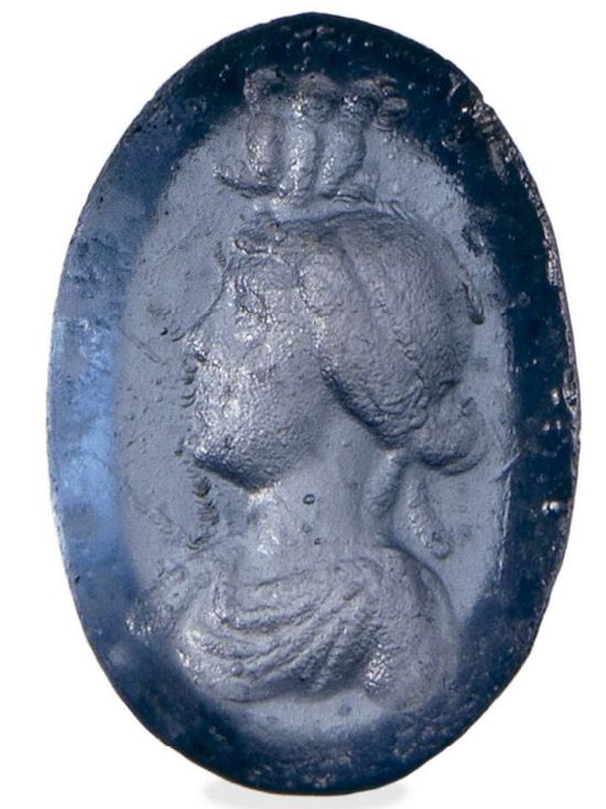 An oval blue glass. It has the portrait of a woman. On top of her head, there is a triple asp. Her hair is pulled back in a chignon. She wears a diadem and Greek clothes. She has a small nose and a pointy chin.