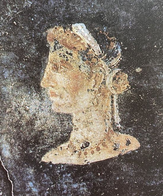 Another painting of a woman in profile. The background is again black. The painting is in bad condition. The woman seems to have reddish-golden hair pulled back in a chignon. She wears a thin band on her head. Her forehead is straight and slants back, her nose is straight and long, her lips are thick, and her chin is rounded. Her face is less long than the previous one. She wears earrings and a golden necklace. Her skin is fair.