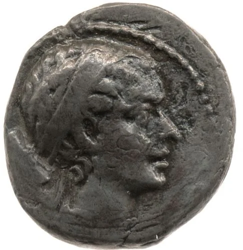 A silver coin with the face of a woman. She is pretty. She has her hair pulled back and wears a diadem. Her forehead is medium-sized and straight, her nose is aquiline. She has full lips and a strong, defined chin.