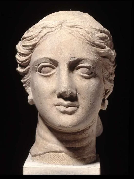 A marble bust of a woman. Her hair is parted in the middle. She has curved eyebrows, wide-set eyes, a straight, long nose, and oval face.