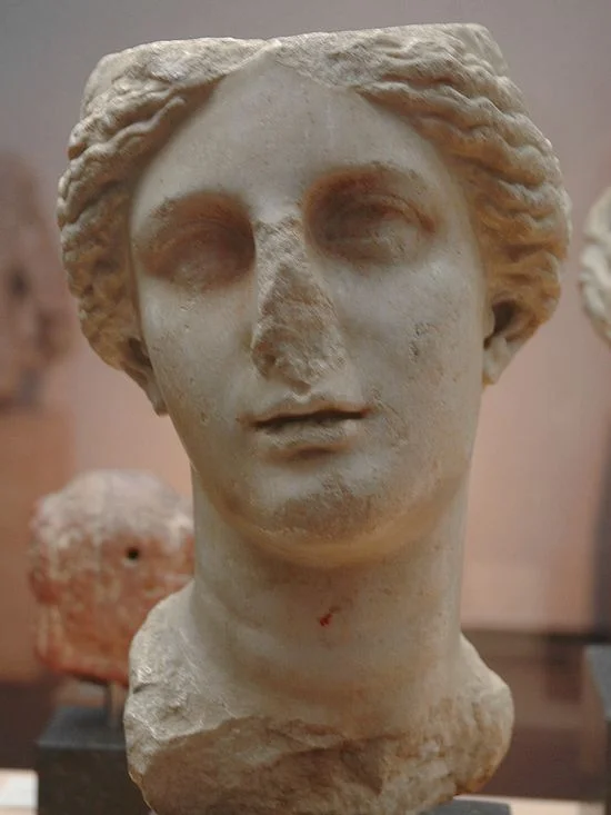 Another marble head of a woman. She, too, has and oval face and hair parted in the middle. Her eyebrows are straighter, her eyes are deep-set, her nose is missing, she has high-cheek bones, a medium-sized mouth and a rounded, strong chin.