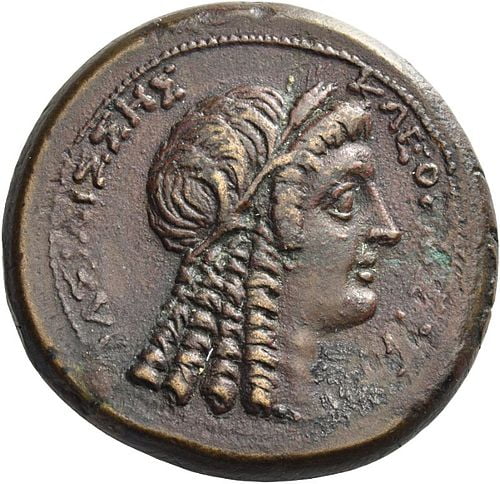 Coin with the face of a owman who wears a ritual, Egyptian wig. She has a stereotypical Greek profile, with a long, straigh nose, and a prominent, rounded chin.