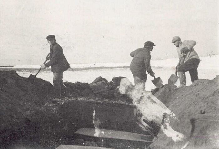 Three men digging a large grave. Inside the grave are some coffins.