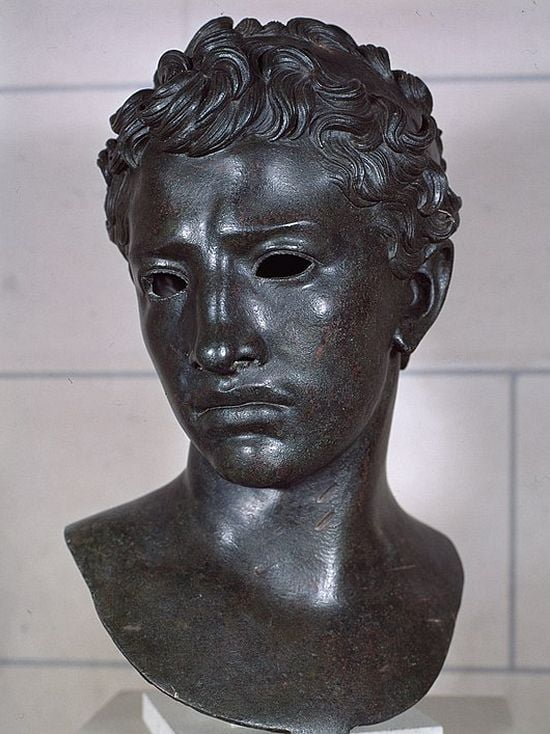 Bronze head of a young man. His hair is wavy and his face is oval-shaped. He has big eyes, a bit wide-set. His nose is broad and somewhat flat, his lips are thick, his chin is round. He is looking down introspectively.