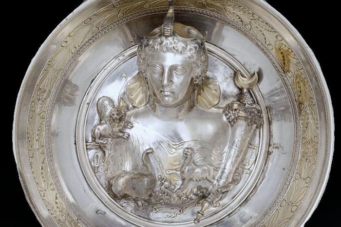 A decorative silver dish. There is a woman sculptured in the middle. She is surrounded by a cornucopia and animals like a lion, cobra, and a panther. The woman wears an elephant headdress, she has wavy hair, a forehead that slants backwards, curved eyebrows, big eyes, an aquiline nose, small mouth, prominent rounded chin. The work is Roman or Greek in style.