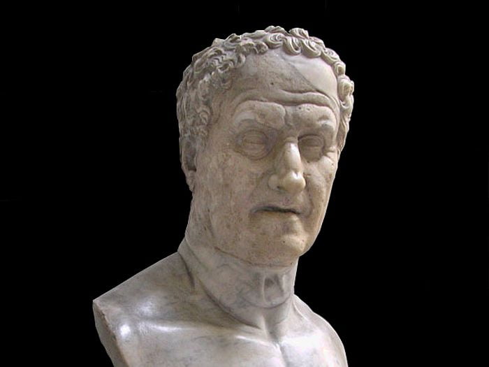 A marble bust of a man. He is probably in his 70s. The statue is expressive, he looks stern. His hair is curly, his forehead broad and with wrinkles. He has a strong Roman nose and deep-set eyes. His face is rectangular, his lips are thin, his jaw is squared. He is knitting his eyebrows in concentration.
