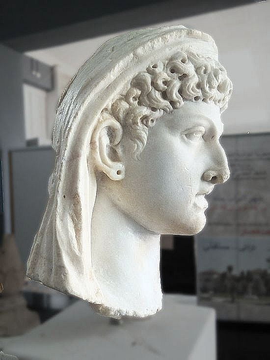 Marble face of a young woman. She wears a veil and has her hair carefully done in curls. She has big ears, a small forehead, a big, aquiline nose, and a prominent chin. Her eyebrows are curved, her eyes deep-set, and she has highish cheekbones. Her neck is thick.