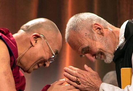 The Dali Lama and another man bow towards each other smiling in a gesture of respect.