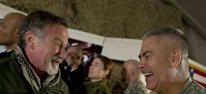 Robin Williams and another man are inside a military base. Robin is saying something to the officer and making him laugh.